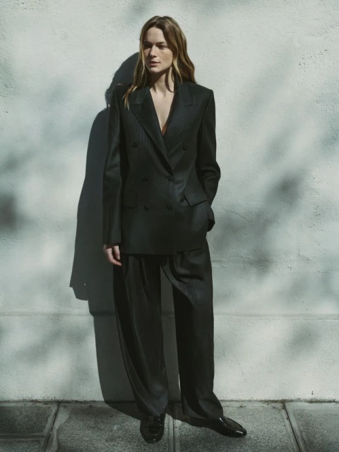 Fashionable woman in a dark pinstripe blazer and pants stands in front of a textured light wall with shadows creating an editorial look.