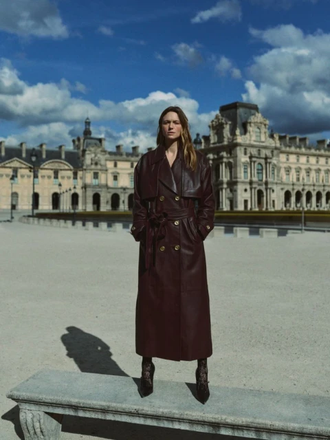 Adult female posing in a maroon trench coat in front of the Louvre on a sunny day. The architectural backdrop complements the fashion.