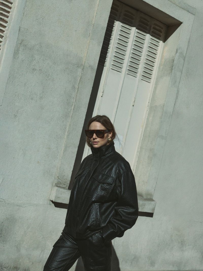 Model wearing a black leather jacket and sunglasses standing outdoors against a light building exterior with a louvered window