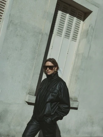 Model wearing a black leather jacket and sunglasses standing outdoors against a light building exterior with a louvered window