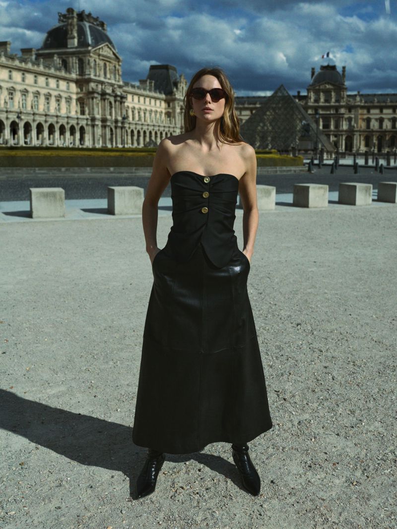 Woman in black dress poses in front of Louvre Museum in Paris. The sky is cloudy and the architectural details are clear.