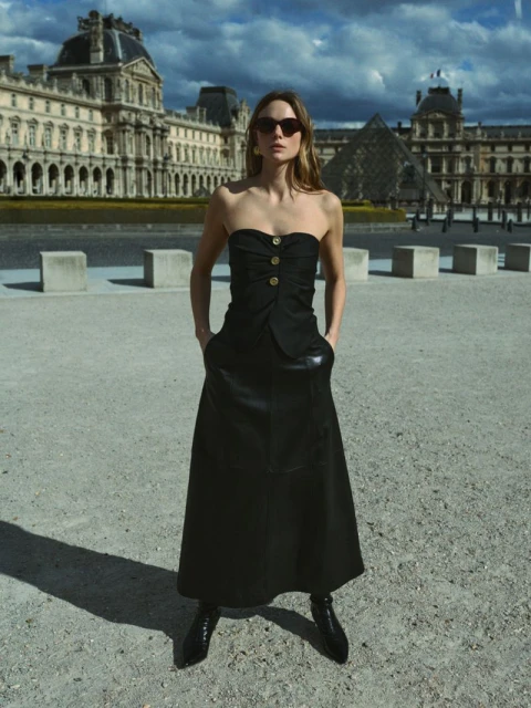 Woman in black dress poses in front of Louvre Museum in Paris. The sky is cloudy and the architectural details are clear.
