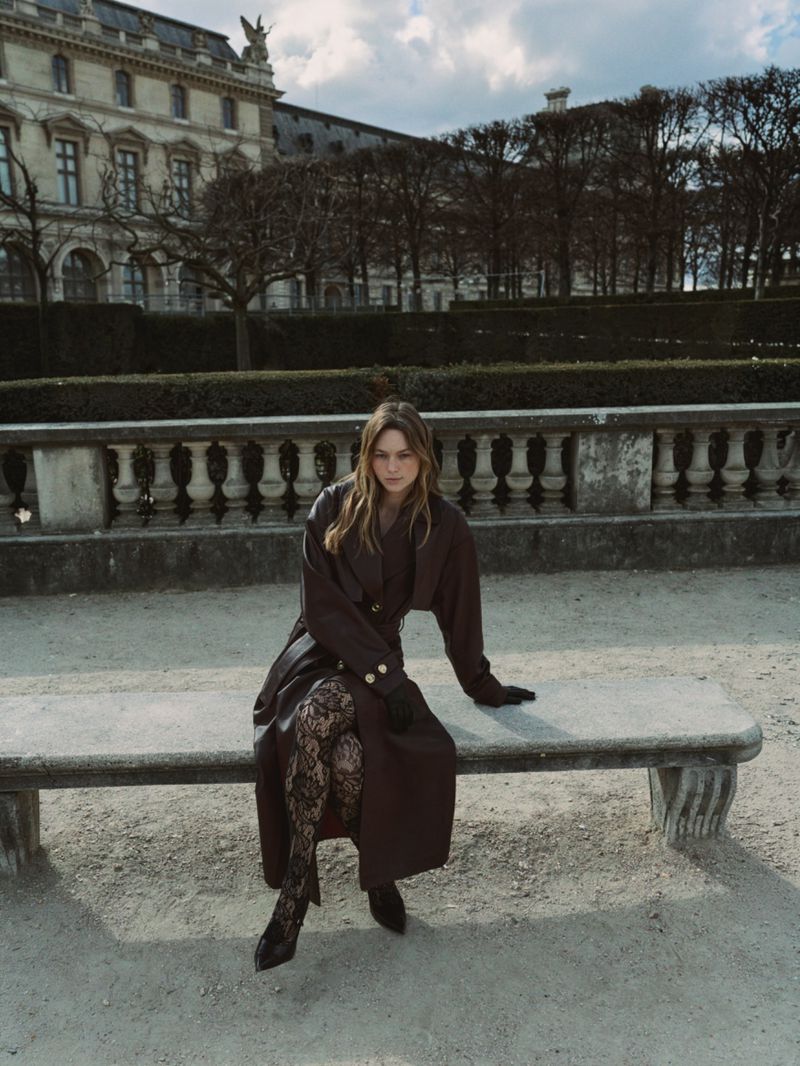 A fashionable young woman in a brown coat and patterned tights sits on a bench outdoors in front of historic architecture and trees.