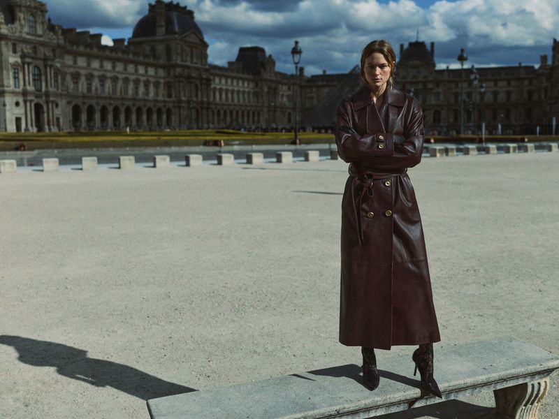 A woman in a long, dark-red leather trench coat poses on a bench in an urban plaza, with a formal French building in the background.