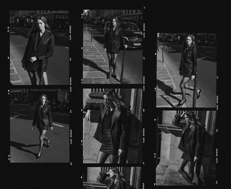Black and white fashion shot of a woman in a leather jacket and skirt walking on a city street with buildings in the background.