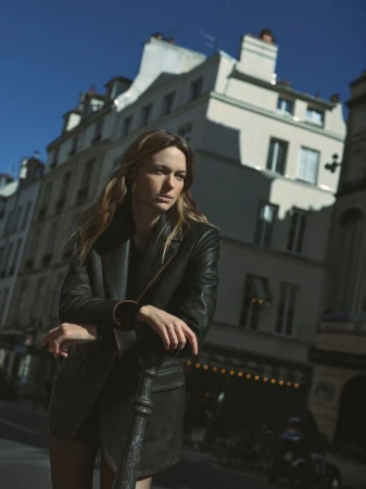 Portrait of a woman in a black blazer and skirt leaning against a metal post, with an urban street and architecture in the background.