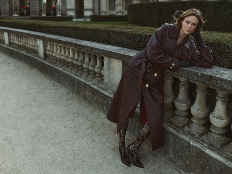 A fashionable woman poses outdoors in a long brown trench coat, lace tights, and heels against a stone balustrade with trimmed hedges.