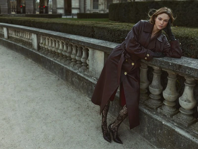 A fashionable woman poses outdoors in a long brown trench coat, lace tights, and heels against a stone balustrade with trimmed hedges.