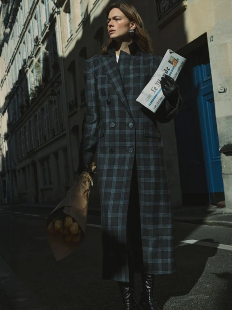 Stylish adult woman in a checkered coat and boots walks down a city street with a newspaper and flowers in her hands.