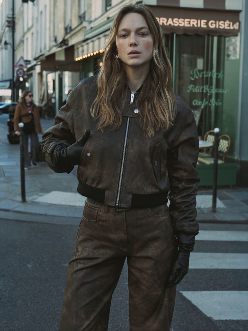 A woman in a brown leather jacket and pants stands on a Paris street near a cafe, captured in a medium shot with natural lighting.