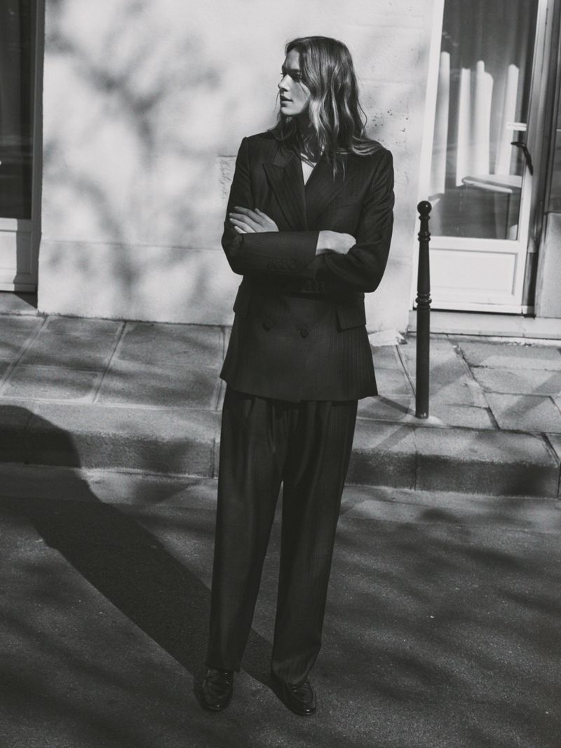 A woman in a dark suit stands on a sidewalk with her arms crossed against a light wall with tree shadows for a fashion shot.