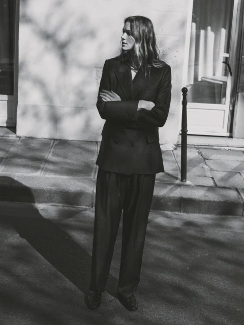 A woman in a dark suit stands on a sidewalk with her arms crossed against a light wall with tree shadows for a fashion shot.
