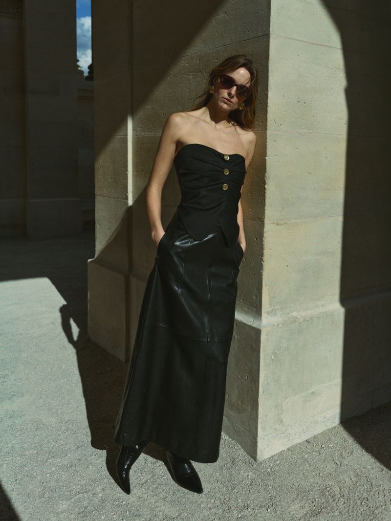 Adult female fashion model in black strapless top and leather skirt, posing in a colonnade with a neutral expression and sunglasses.