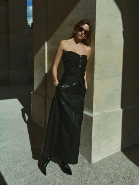 Adult female fashion model in black strapless top and leather skirt, posing in a colonnade with a neutral expression and sunglasses.