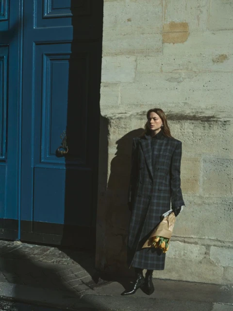 A young woman in a plaid coat stands outside a building with a blue door, holding a paper bag with yellow flowers in warm light.