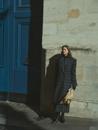 Model in a plaid coat holding a paper-wrapped bouquet standing before a blue double door and a stone wall.