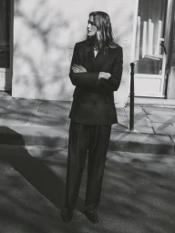 Model standing outdoors with arms crossed wearing a dark blazer and trousers against a building wall