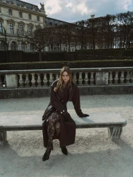 Model seated on a stone bench wearing a long dark coat and patterned tights outdoors against a classic building