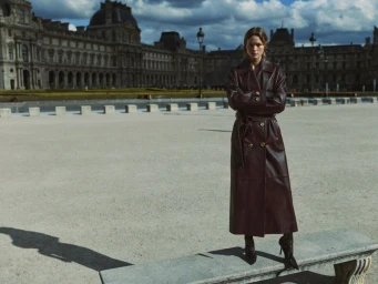 Model wearing a long burgundy leather coat standing outdoors in front of a classical European palace building