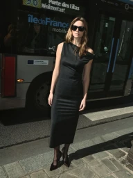 Model wearing dark sunglasses and a black dress standing on a stone pavement in front of a bus