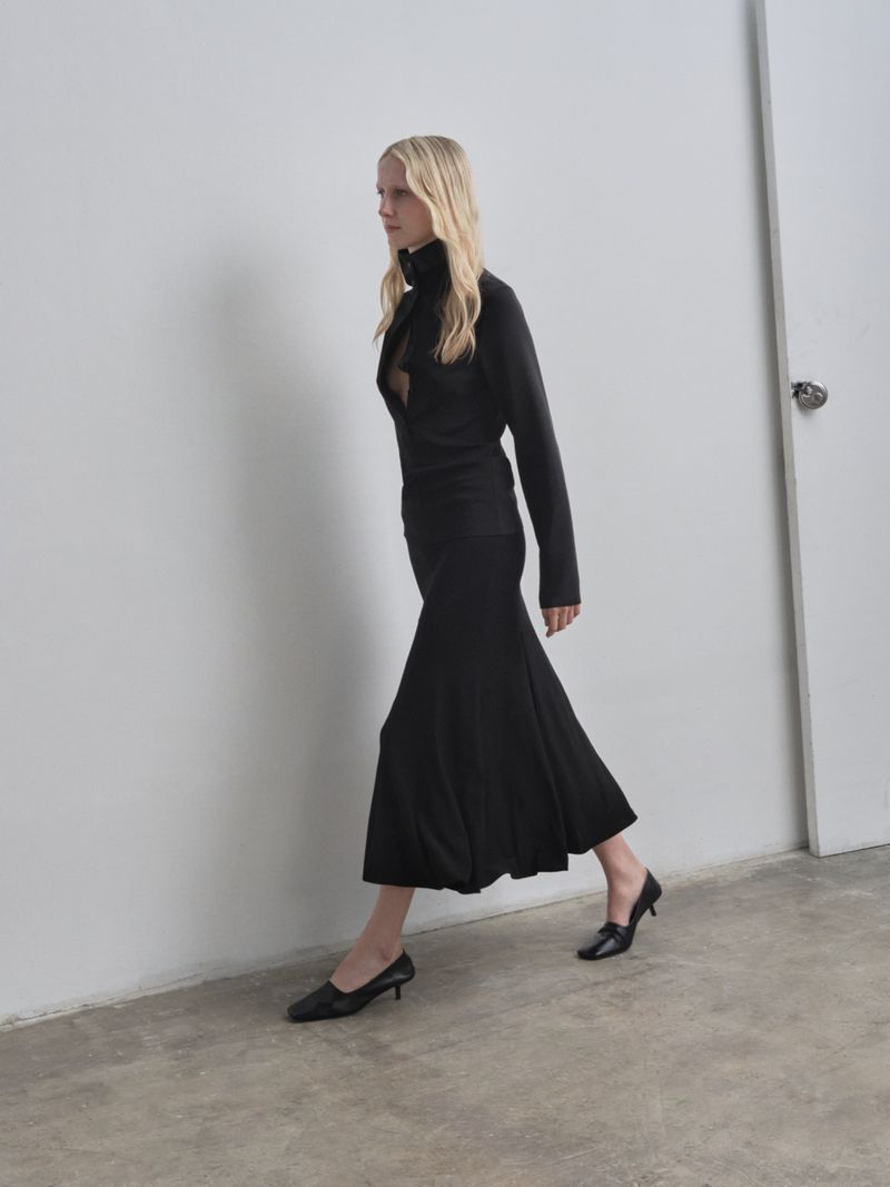 A blonde woman in a black, long-sleeved top and midi skirt walks in profile against a white wall, showcasing fashion and simple indoor setting.