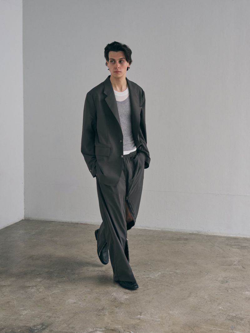 Young adult in a grey suit and white t-shirt stands in a minimalist room. The outfit is oversized, and the person is looking to the side.