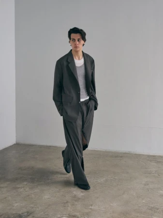 Young adult in a grey suit and white t-shirt stands in a minimalist room. The outfit is oversized, and the person is looking to the side.