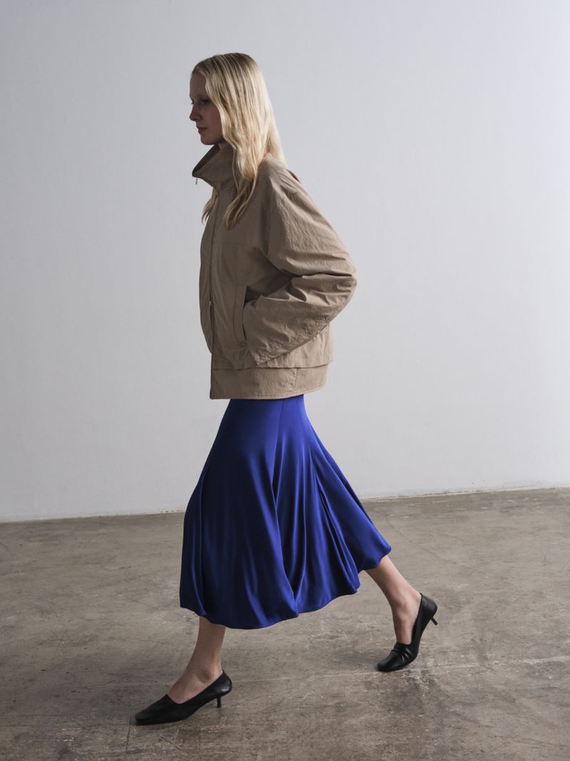 A woman walks in a studio, wearing a tan jacket and a blue skirt with black heels. The background is a solid white wall and gray floor.