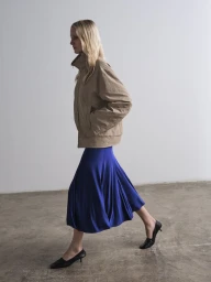 Model in profile wearing a tan jacket and blue skirt against a neutral studio backdrop