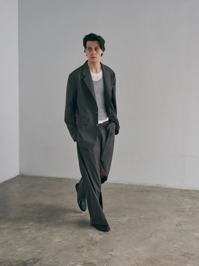 A young man wearing a dark grey suit and layered tops walks across a concrete floor against a plain grey wall.