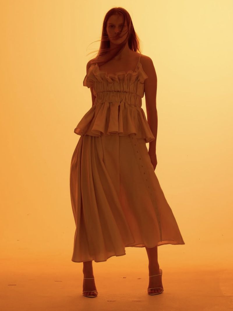 A woman stands in a photo studio wearing a light-colored, two-piece dress and heels, against a plain orange backdrop, full-length view.