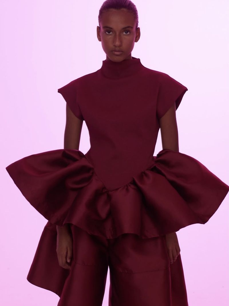 Fashion model poses in a sleeveless, high-necked dark red top with large ruffles and matching culottes against a pink background.