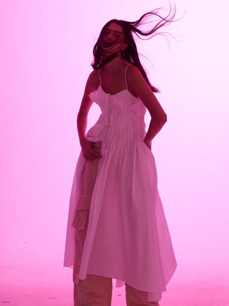 Woman in a flowing white dress stands against a bright pink background with her hair blowing in the wind in a fashion forward photo.