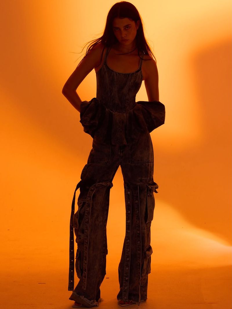 Woman with long hair poses in a denim jumpsuit with pockets, set against a warm orange background, showcasing a fashion-forward studio shot.