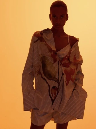 Fashion shot of a woman in oversized striped shirt and shorts in a studio with yellow lighting, posing for the camera with a neutral expression.