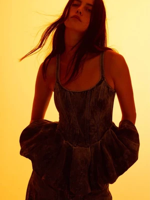 Model Ariana Soeiro wearing a dark denim top with a flared peplum waist against a warm golden gradient background.