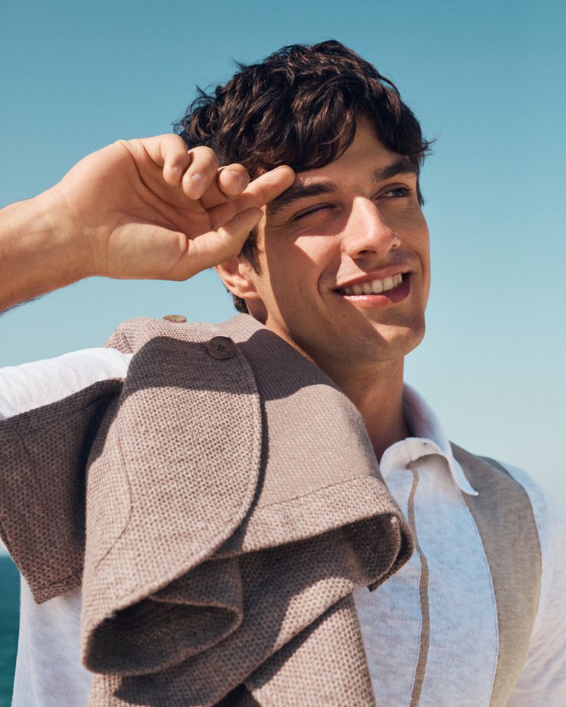 Smiling man wearing a white and taupe polo shirt, standing outside against a clear sky with a woven jacket draped over his shoulder.
