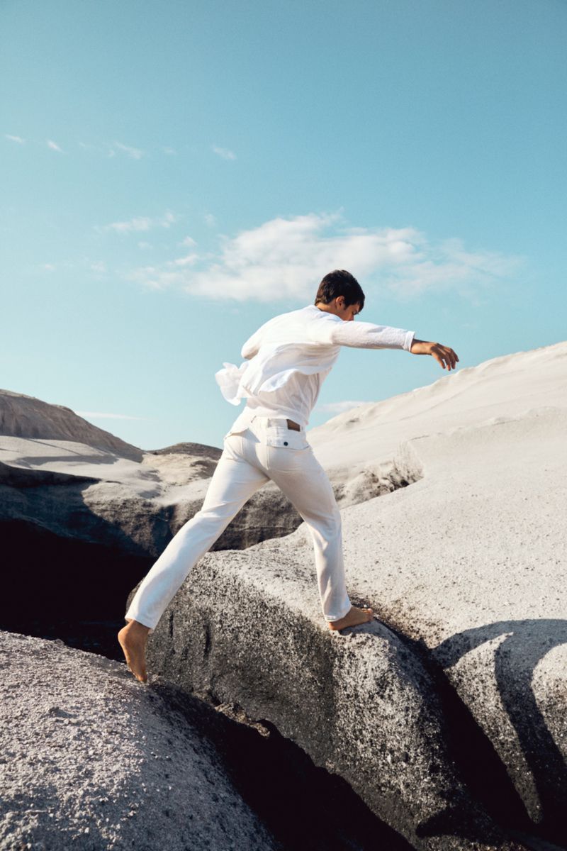 Man in white apparel leaps across rock formation under a blue sky. Barefoot on rock, clothing in motion, in outdoor landscape.