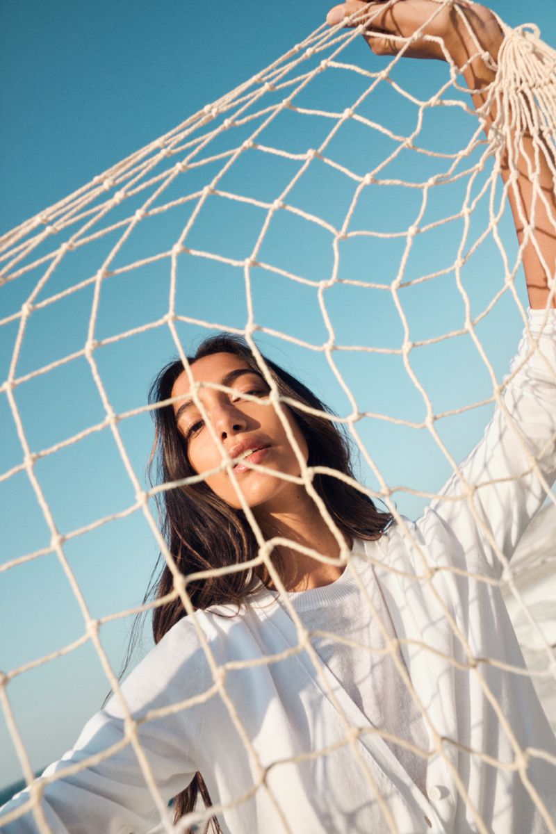 Portrait of a woman with long hair in a white top looking through a white net against a blue sky on a sunny day.