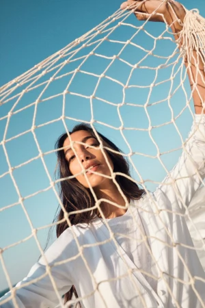 Portrait of a woman with long hair in a white top looking through a white net against a blue sky on a sunny day.