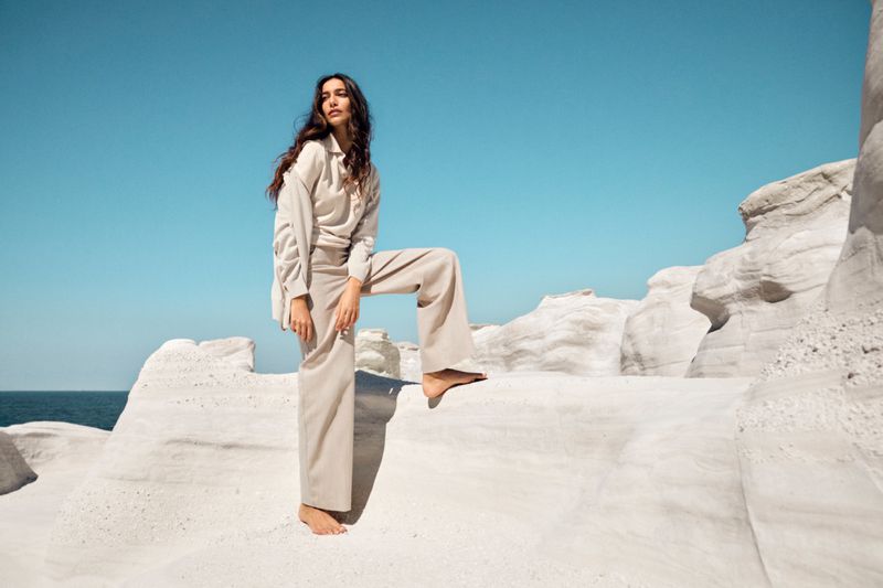 A woman in beige clothing poses on a rocky, white landscape under a clear blue sky, standing barefoot with one leg up.