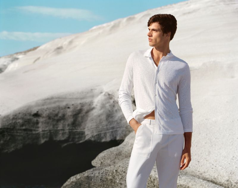 A male fashion model poses outdoors in a white long-sleeved shirt and pants with a blurred mountain landscape backdrop under a clear sky.