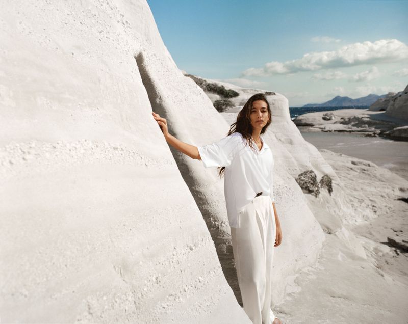 A woman in white clothing poses against a white rock formation on a sunny day near the ocean, exuding a serene and stylish aesthetic.