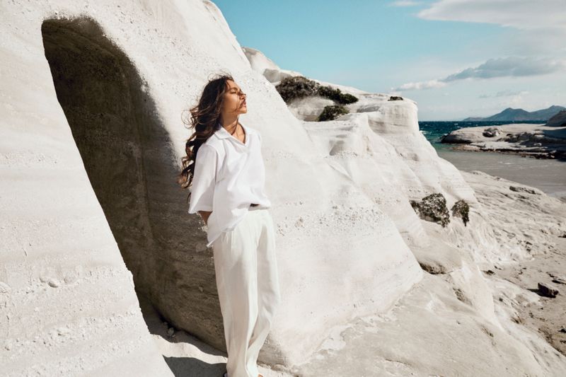 Woman in white clothing stands in a natural stone cave on a sunny day by the sea, gazing at the sky with her eyes closed.