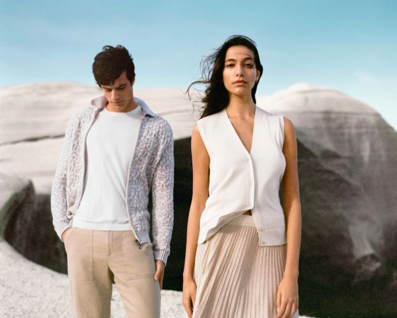 A stylish couple poses in front of a rock formation under a light blue sky. The man wears a cardigan, t-shirt, and pants; the woman wears a sweater vest and skirt.