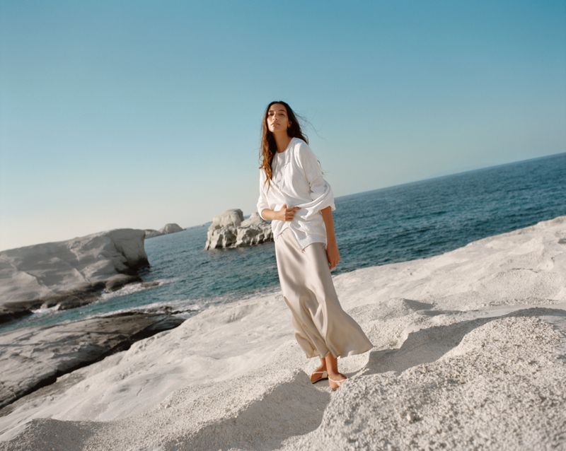 Adult female standing on white rocky shore near blue ocean water and sky on a sunny day in an outdoor environment.