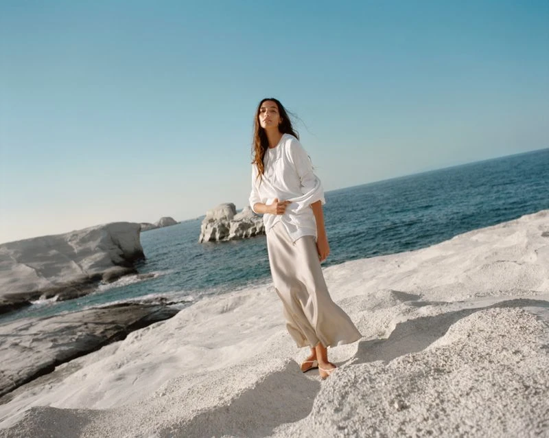 Adult female standing on white rocky shore near blue ocean water and sky on a sunny day in an outdoor environment.