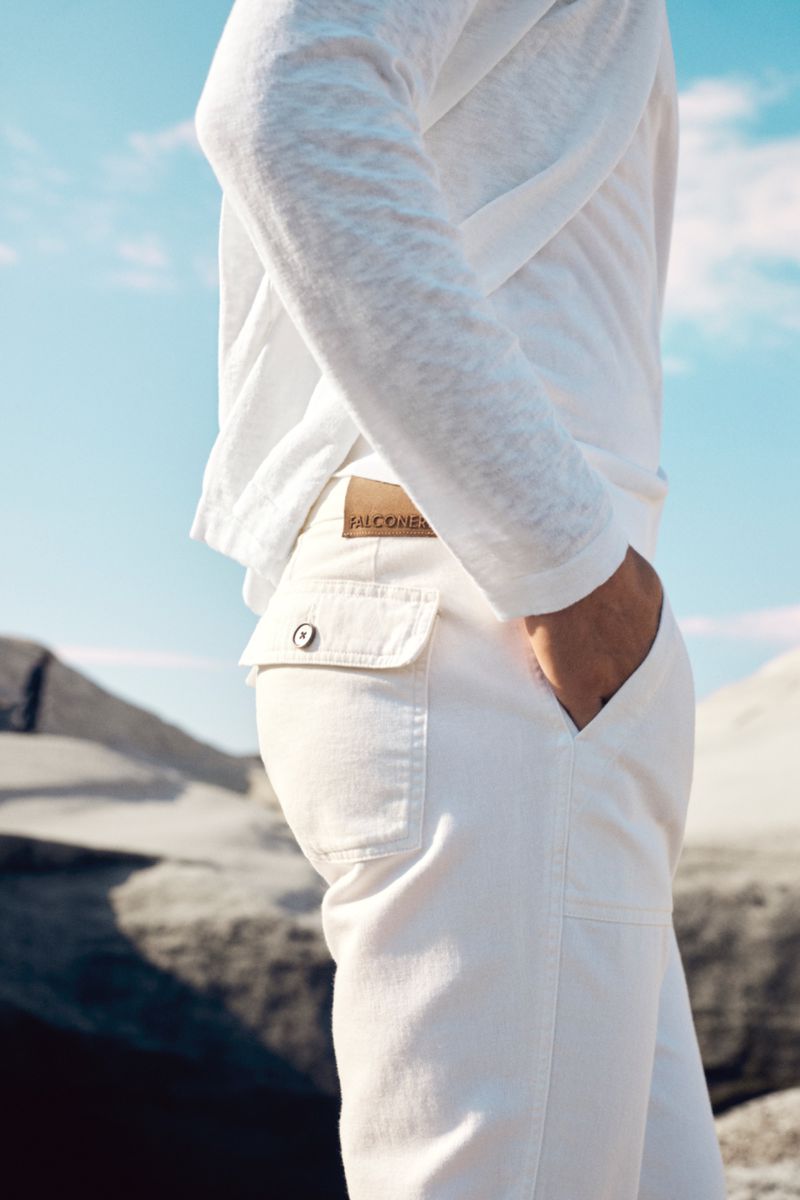 Fashion shot of a man's lower torso wearing white pants and a long-sleeved white shirt with a mountain background and bright sky.