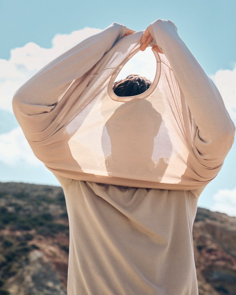 Person pulls a beige t-shirt over their head outdoors against a sky with clouds and a hilly landscape in the background.