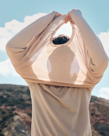 Person pulls a beige t-shirt over their head outdoors against a sky with clouds and a hilly landscape in the background.